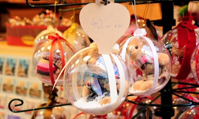 Holiday-themed ornaments displayed in a market. Each ornament contains a small teddy bear and artificial snow inside a clear plastic sphere. They are adorned with colorful ribbons, and a heart-shaped tag with pricing information is attached to one of the ornaments.