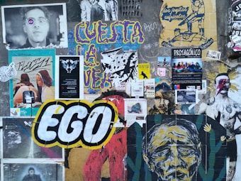 A collage of various posters and graffiti covers a wall. The large word 'EGO' is prominently featured in bold white letters with a black and yellow outline. Surrounding it are multiple artworks, including portraits, abstract designs, and text. There are images of people, some with artistic alterations such as added colors and patterns on their faces. The overall composition is colorful and layered, reflecting an eclectic mix of styles and messages.