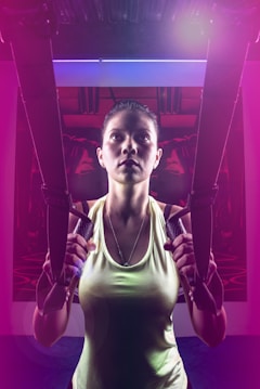 A person is using suspension trainers in a fitness environment. The lighting is dramatic, with a mix of bright and dark areas, creating a focused and intense atmosphere. The image captures the person's concentration and physical effort.