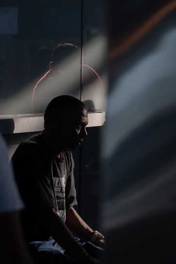A person sits in shadow, possibly in deep thought or introspection. The lighting is dramatic, with a beam of light cutting across the scene, highlighting part of the individual's profile. A reflective surface behind them creates a faint reflection.