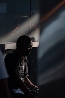 A person sits in shadow, possibly in deep thought or introspection. The lighting is dramatic, with a beam of light cutting across the scene, highlighting part of the individual's profile. A reflective surface behind them creates a faint reflection.