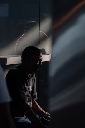 A person sits in shadow, possibly in deep thought or introspection. The lighting is dramatic, with a beam of light cutting across the scene, highlighting part of the individual's profile. A reflective surface behind them creates a faint reflection.