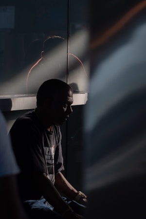 A person sits in shadow, possibly in deep thought or introspection. The lighting is dramatic, with a beam of light cutting across the scene, highlighting part of the individual's profile. A reflective surface behind them creates a faint reflection.