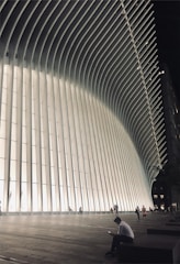 A futuristic building with a series of tall, vertical white columns that curve outward towards the top. The structure is illuminated, creating a contrast with the darker surroundings. Several people are visible walking or standing near the structure, with one person sitting and looking at a device in the foreground.