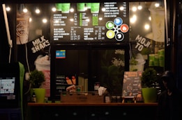 A small coffee shop counter with a menu board displaying various coffee drinks and prices in both traditional and metric units. Bright lights illuminate the menu, and there are green cups stacked beside the counter. Potted plants decorate the counter along with promotional posters for milkshakes, hot dogs, and paninis. There are also signs highlighting different coffee options and flavors.
