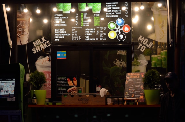 A small coffee shop counter with a menu board displaying various coffee drinks and prices in both traditional and metric units. Bright lights illuminate the menu, and there are green cups stacked beside the counter. Potted plants decorate the counter along with promotional posters for milkshakes, hot dogs, and paninis. There are also signs highlighting different coffee options and flavors.