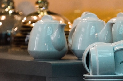 White ceramic teapots and cups are neatly arranged on a table, with reflective surfaces creating soft highlights. The background showcases blurred, warm lighting from what appear to be glass objects or decor.