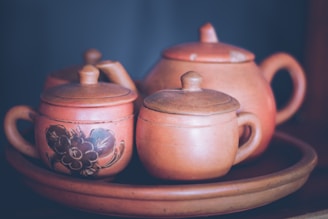 A collection of clay teapots and cups with lids resting on a matching clay tray. One of the teapots features a floral pattern in black. The set has a rustic, handcrafted appearance with earthy tones.