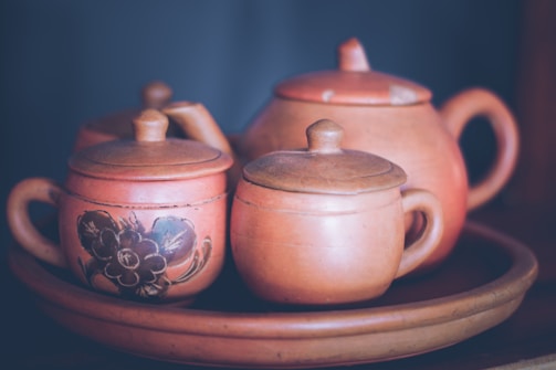 A collection of clay teapots and cups with lids resting on a matching clay tray. One of the teapots features a floral pattern in black. The set has a rustic, handcrafted appearance with earthy tones.
