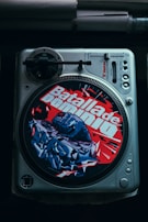 A vintage vinyl turntable with a colorful slipmat featuring bold red and blue graffiti art and text. The slipmat displays the words 'Batalla de Dominio' with an image of a fist and dynamic shapes. The turntable includes control knobs and a tonearm, suggesting a focus on DJing or music.