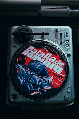 A vintage vinyl turntable with a colorful slipmat featuring bold red and blue graffiti art and text. The slipmat displays the words 'Batalla de Dominio' with an image of a fist and dynamic shapes. The turntable includes control knobs and a tonearm, suggesting a focus on DJing or music.