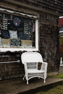 A white wicker chair is placed outside a weathered wooden building with a shingle exterior. The window of the building displays various decorative items, including pillows with nautical designs, hanging beads, and a sign with the word 'Maine'. A small sign in the window describes the shop as having unique gift ideas with interesting stories. The overall atmosphere appears quaint and rustic.