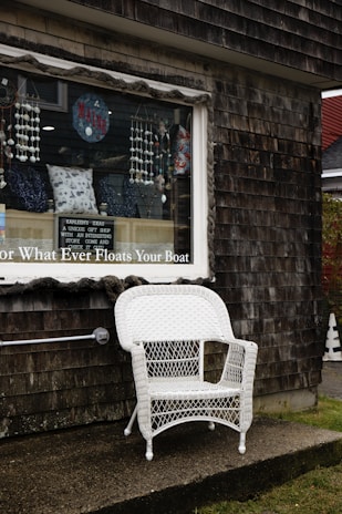 A white wicker chair is placed outside a weathered wooden building with a shingle exterior. The window of the building displays various decorative items, including pillows with nautical designs, hanging beads, and a sign with the word 'Maine'. A small sign in the window describes the shop as having unique gift ideas with interesting stories. The overall atmosphere appears quaint and rustic.