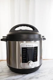 A stainless steel electric pressure cooker positioned on a marble surface with a digital display on the front panel. The cooker has a black lid with a handle and various function buttons.