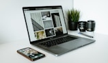 A sleek laptop displaying a series of black and white images sits on a white desk. Next to the laptop are two camera lenses, a pen, a smartphone with various apps visible on its screen, and a small potted plant with vibrant green leaves.