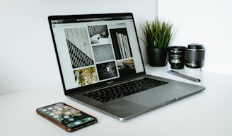 A sleek laptop displaying a series of black and white images sits on a white desk. Next to the laptop are two camera lenses, a pen, a smartphone with various apps visible on its screen, and a small potted plant with vibrant green leaves.