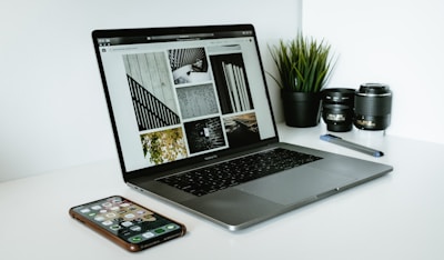 A sleek laptop displaying a series of black and white images sits on a white desk. Next to the laptop are two camera lenses, a pen, a smartphone with various apps visible on its screen, and a small potted plant with vibrant green leaves.