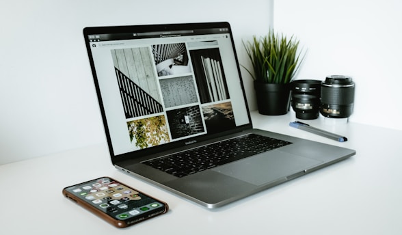 A sleek laptop displaying a series of black and white images sits on a white desk. Next to the laptop are two camera lenses, a pen, a smartphone with various apps visible on its screen, and a small potted plant with vibrant green leaves.