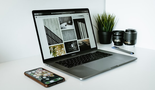 A sleek laptop displaying a series of black and white images sits on a white desk. Next to the laptop are two camera lenses, a pen, a smartphone with various apps visible on its screen, and a small potted plant with vibrant green leaves.
