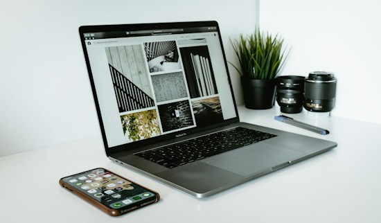 A sleek laptop displaying a series of black and white images sits on a white desk. Next to the laptop are two camera lenses, a pen, a smartphone with various apps visible on its screen, and a small potted plant with vibrant green leaves.