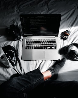 MacBook in middle of camera, action camera, mask, and headphones on bed