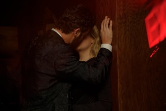 Two people, a man and a woman, are intimately embracing against a wooden wall in a dimly lit environment. The man is wearing a dark textured suit, and the woman's face is partially obscured by the man's arm. The lighting casts a warm glow, and there is a red exit sign reflected in the background.