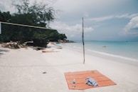 A tranquil beach scene with a volleyball net set up on white sand. Nearby, an orange beach mat with a water bottle and a piece of cloth is laid out. In the distance, lush green trees and rocks border the shoreline. The blue ocean stretches out towards the horizon under a partly cloudy sky, with a few people swimming.