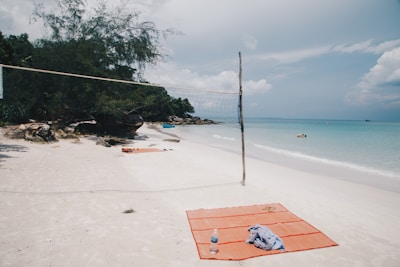 A tranquil beach scene with a volleyball net set up on white sand. Nearby, an orange beach mat with a water bottle and a piece of cloth is laid out. In the distance, lush green trees and rocks border the shoreline. The blue ocean stretches out towards the horizon under a partly cloudy sky, with a few people swimming.