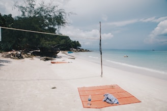 A tranquil beach scene with a volleyball net set up on white sand. Nearby, an orange beach mat with a water bottle and a piece of cloth is laid out. In the distance, lush green trees and rocks border the shoreline. The blue ocean stretches out towards the horizon under a partly cloudy sky, with a few people swimming.