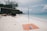 A tranquil beach scene with a volleyball net set up on white sand. Nearby, an orange beach mat with a water bottle and a piece of cloth is laid out. In the distance, lush green trees and rocks border the shoreline. The blue ocean stretches out towards the horizon under a partly cloudy sky, with a few people swimming.
