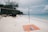A tranquil beach scene with a volleyball net set up on white sand. Nearby, an orange beach mat with a water bottle and a piece of cloth is laid out. In the distance, lush green trees and rocks border the shoreline. The blue ocean stretches out towards the horizon under a partly cloudy sky, with a few people swimming.
