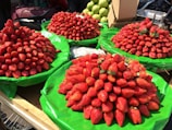 A market stall displays several large piles of fresh strawberries on green tarps. The strawberries are vibrant red and vary slightly in shape and size. In the background, there are green fruits that appear to be guavas and a cardboard box.