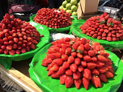A market stall displays several large piles of fresh strawberries on green tarps. The strawberries are vibrant red and vary slightly in shape and size. In the background, there are green fruits that appear to be guavas and a cardboard box.
