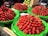 A market stall displays several large piles of fresh strawberries on green tarps. The strawberries are vibrant red and vary slightly in shape and size. In the background, there are green fruits that appear to be guavas and a cardboard box.