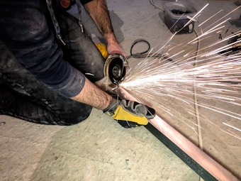 A person wearing protective gloves is using an angle grinder to cut through a metal rod on a concrete floor. Sparks are flying outwards from the point of contact, creating a dynamic visual effect. The individual is kneeling and appears focused on the task.
