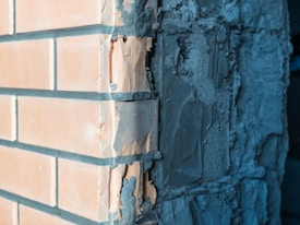 A close-up view of a corner between two walls, one covered with unfinished plaster and the other with brickwork. The plaster appears rough and uneven, with visible textures and imperfections. The bricks are neatly arranged, displaying a light orange color.
