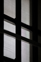 A close-up view of a window with a mesh screen, creating a grid-like pattern that contrasts with the light coming through the glass panes.