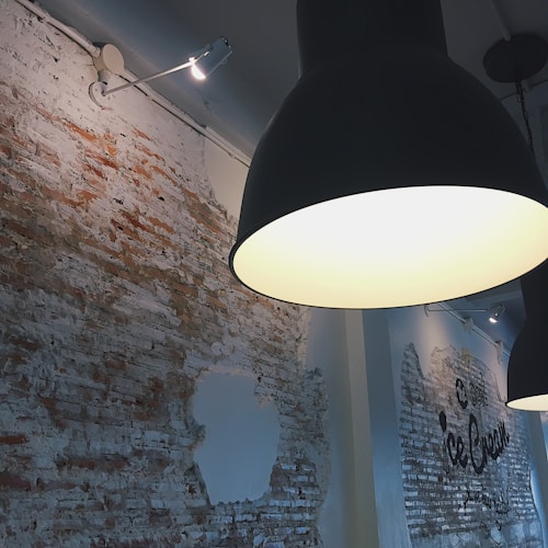 The image captures an industrial-style interior with exposed brick walls partially covered with white paint. A prominent black pendant light hangs from the ceiling, casting a warm glow. Overhead track lighting highlights sections of the brickwork where a stencil design is partially visible.