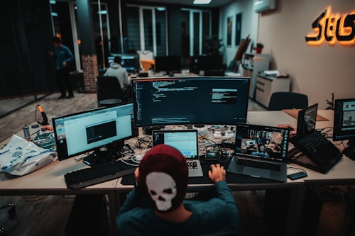 A person wearing a cap with a skull design sits at a cluttered desk in a dimly lit office. Multiple computer monitors display code and web pages, suggesting a tech-related work environment. Wires and devices are scattered across the desk, indicating a busy or engaged workspace. In the background, other individuals are visible, contributing to an active and collaborative atmosphere.