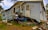 A significantly damaged house with its structure leaning precariously and a partially crushed blue vehicle wedged underneath. The house's siding is warped and twisted, with debris scattered around.