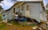 A significantly damaged house with its structure leaning precariously and a partially crushed blue vehicle wedged underneath. The house's siding is warped and twisted, with debris scattered around.