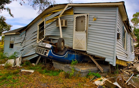 A significantly damaged house with its structure leaning precariously and a partially crushed blue vehicle wedged underneath. The house's siding is warped and twisted, with debris scattered around.