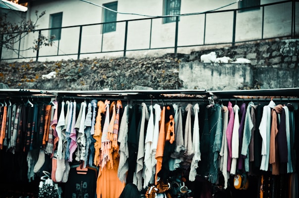 A variety of colorful scarves and garments are hanging on a rack, displayed outside, possibly in a market setting. The background includes a rough stone wall and a building with small windows.