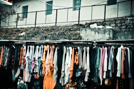 A variety of colorful scarves and garments are hanging on a rack, displayed outside, possibly in a market setting. The background includes a rough stone wall and a building with small windows.