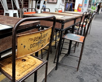 Several worn-out wooden and metal school desks and chairs arranged in a row. The backrest of one chair is rusty and has some faded writing on it. The setting appears to be outdoors, likely on a paved surface. Some other chairs in the background have white frames and are stacked together.
