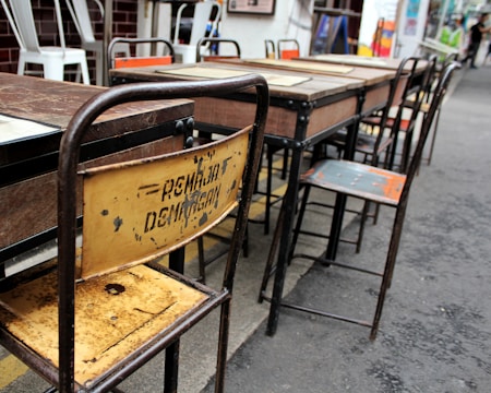 Several worn-out wooden and metal school desks and chairs arranged in a row. The backrest of one chair is rusty and has some faded writing on it. The setting appears to be outdoors, likely on a paved surface. Some other chairs in the background have white frames and are stacked together.