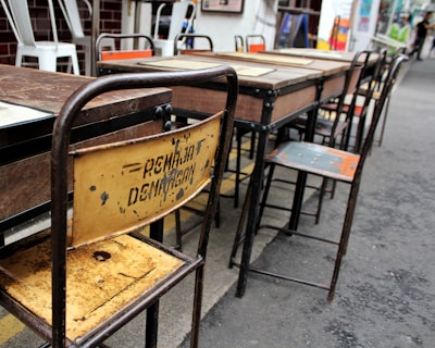 Several worn-out wooden and metal school desks and chairs arranged in a row. The backrest of one chair is rusty and has some faded writing on it. The setting appears to be outdoors, likely on a paved surface. Some other chairs in the background have white frames and are stacked together.