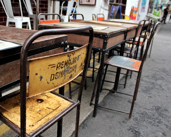 Several worn-out wooden and metal school desks and chairs arranged in a row. The backrest of one chair is rusty and has some faded writing on it. The setting appears to be outdoors, likely on a paved surface. Some other chairs in the background have white frames and are stacked together.