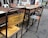 Several worn-out wooden and metal school desks and chairs arranged in a row. The backrest of one chair is rusty and has some faded writing on it. The setting appears to be outdoors, likely on a paved surface. Some other chairs in the background have white frames and are stacked together.
