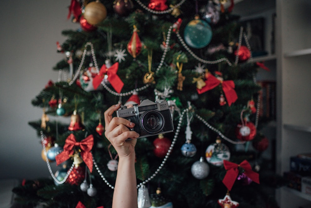 persona che tiene la macchina fotografica nera point-and-shoot vicino all'albero di Natale